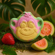 Toy monkey head with pink and green colors surrounded by a strawberry and guava on a leafy background