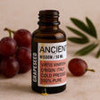 Bottle of Ancient Wisdom grapeseed oil with grapes and an olive leaf on a beige background