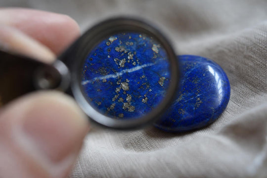 Uma lupa a ampliar um cabochão de lápis-lazúli polido, mostrando uma base azul, manchas douradas de pirite e um veio branco de calcite