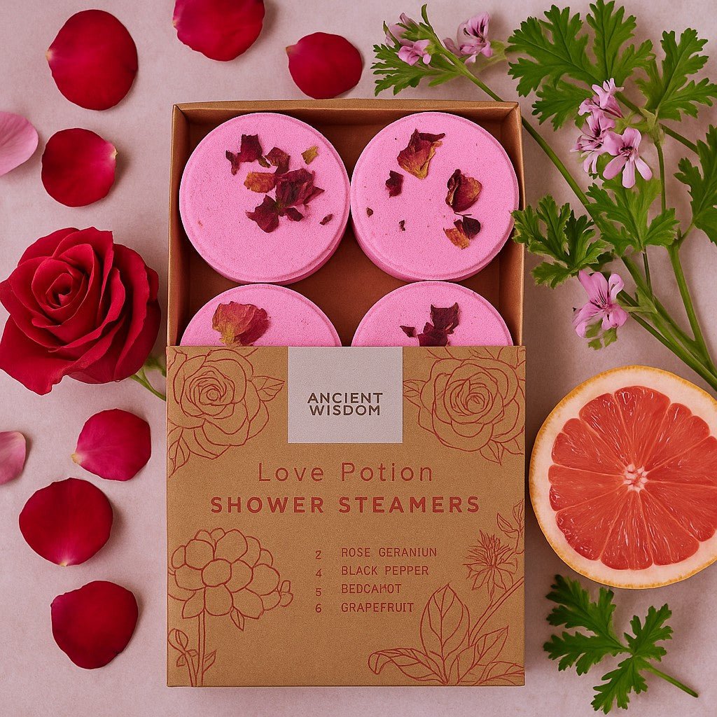 Box of 'Love Potion' shower steamers with pink bath bombs, surrounded by flowers and a grapefruit slice on a pink background.