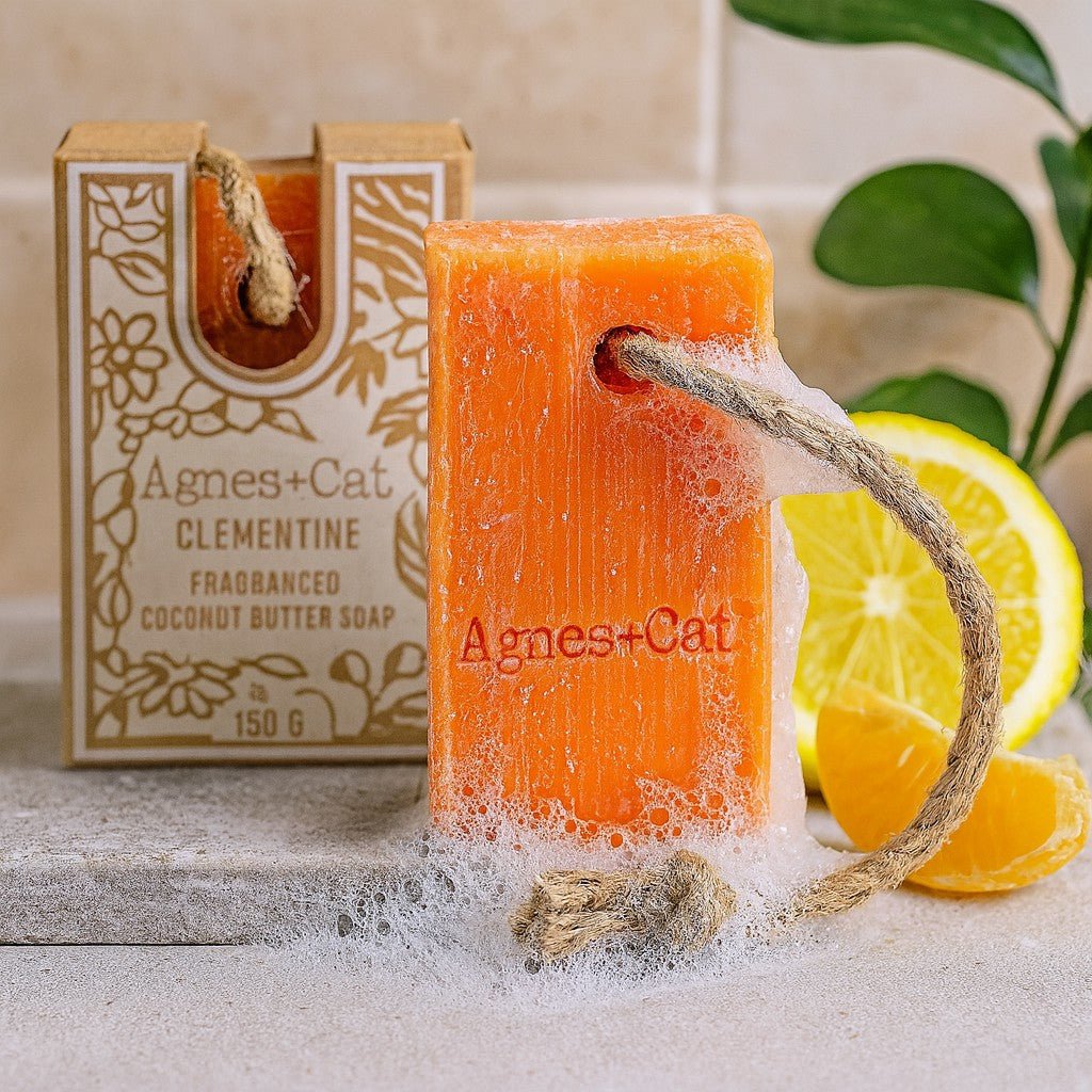 Orange soap bar with a rope, next to its packaging and citrus fruits on a neutral background