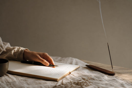 Une main posée sur un stylo, sur un carnet ouvert, avec un mince filet de fumée d'encens qui s'élève à côté.