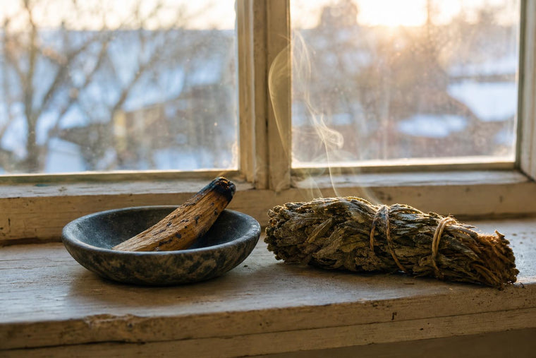 A stick of palo santo wood and a bundle of dried white sage smouldering together in a stone dish on a European windowsill