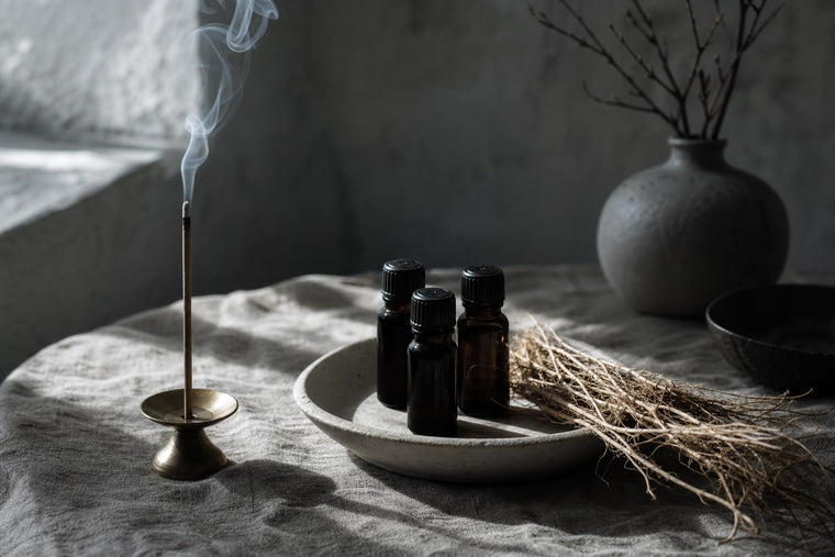 Three dark glass essential oil bottles beside dried vetiver root and a burning frankincense incense stick on unbleached linen, cool north light