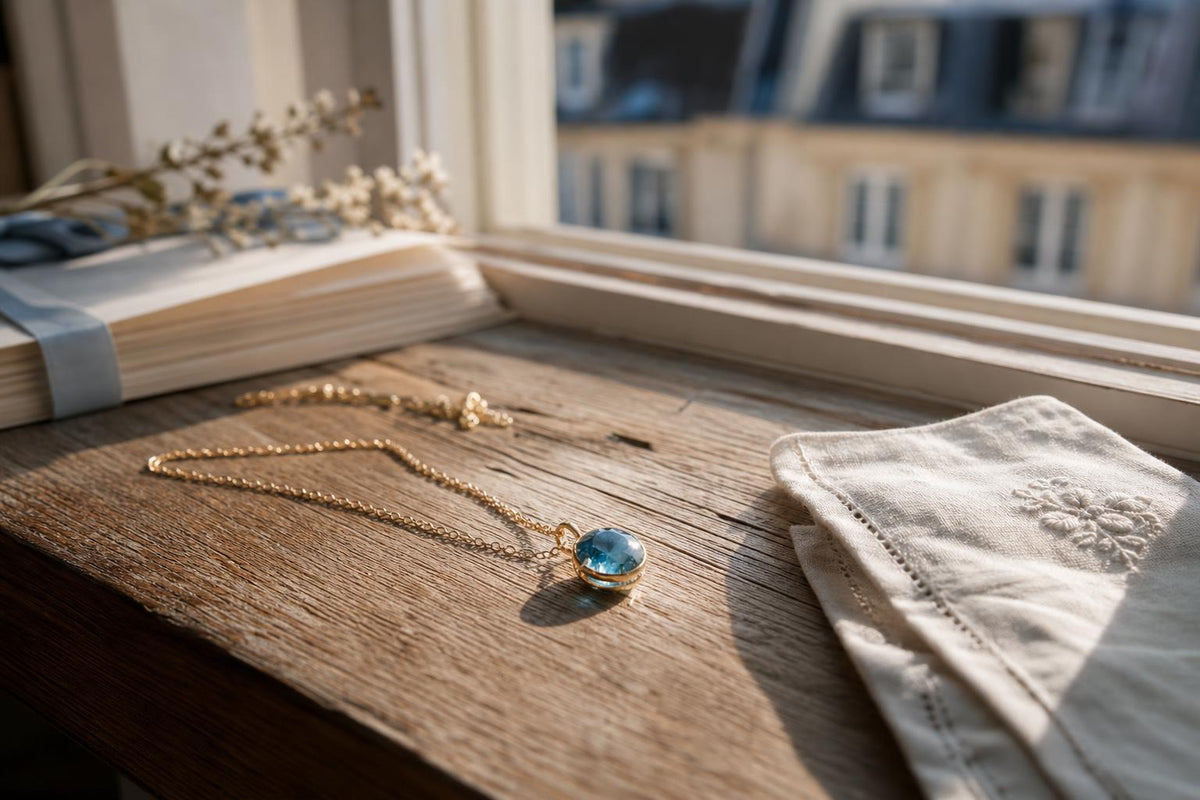 A blue gemstone pendant resting in morning light on a windowsill, beside a folded linen handkerchief and wedding invitations