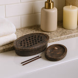 Wooden incense burner with two sticks on a bathroom counter, next to a soap dispenser and candle.