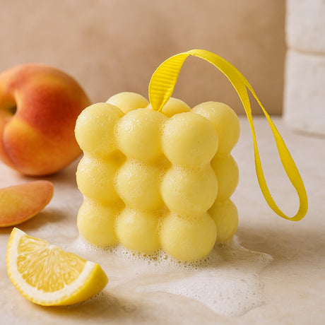 Yellow bath sponge with a handle next to a peach and lemon wedge on a beige surface