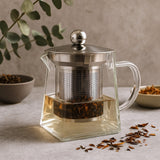 Clear glass teapot with metal lid and tea leaves on a neutral background