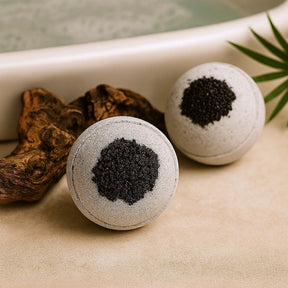 Two bath bombs with black powder on a beige surface with a bathtub in the background.