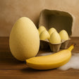 Large yellow egg next to a bunch of bananas on a wooden surface with a cardboard egg carton in the background.