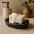 Bar of soap with suds in a black soap dish on a wooden surface, with a gold pump dispenser and a vase in the background.