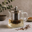 Clear glass teapot with metal lid and tea leaves on a neutral background