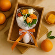 Bouquet of flowers wrapped in paper with an orange ribbon, placed inside a cardboard box on a beige surface with oranges and leaves.