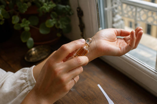Applying perfume to the wrist beside a paper scent-testing strip in natural window light