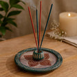 Incense stick holder with incense sticks on a wooden surface, candle, and plant in the background