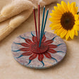 Decorative incense holder with three sticks on a sun-shaped base, next to a sunflower and dried plants.