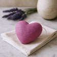 Heart-shaped pink sponge on a white cloth with lavender and a ceramic item in the background.