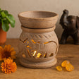 Decorative terracotta oil burner with elephant design on a wooden surface with flowers and a small statue.