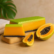 Papaya slices and a green and orange scrubber on a wooden surface with a plant in the background.