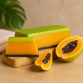 Papaya slices and a green and orange scrubber on a wooden surface with a plant in the background.