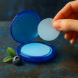 Hand holding a blue circular container with a white disc, next to a blueberry and mint leaves on a dark surface.