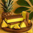 Two bars of soap, one yellow and one brown, on a wooden board with pineapple slices and a whole pineapple in the background.