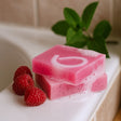 Pink soap with a brand logo on a white surface with raspberries and green leaves.