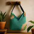 Green and beige handbag with a tag hanging on a wooden hook against a beige wall.