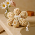 Knitted beige clover-shaped item with a tassel on a wooden surface with flowers and soap in the background.