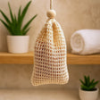 Woven soap bag hanging on a wooden shelf with plants and a towel in the background.