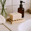 Bar of soap on a wooden soap dish with a bottle and plant in the background