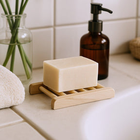 Bar of soap on a wooden soap dish with a bottle and plant in the background