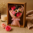 Bouquet of pink and red roses in a box with a candle and additional flowers on a beige surface.