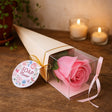 Pink soap flower in a clear box on a wooden surface with candles and a plant in the background.