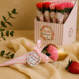 Bouquet of pink flowers wrapped in gold paper with a pink ribbon, placed on a beige surface with a box of similar products in the background.