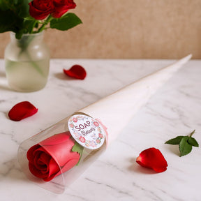 Red rose in a clear packaging with a decorative label on a marble surface with scattered rose petals.