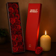 Red roses in a box with a red packaging box labeled 'Merci Bouquet' next to a lit candle on a wooden surface.