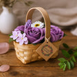 Basket of purple and white flowers with a wooden handle on a wooden surface, featuring a 'Bouq Flowers' tag.