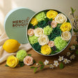 Colorful flower arrangement in a round container with 'Merci Bouquet' branding, surrounded by lemons and flowers on a wooden surface.