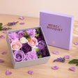 Box of purple and pink flowers with 'Merci Bouquet' packaging on a wooden surface.