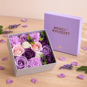 Box of purple and pink flowers with 'Merci Bouquet' packaging on a wooden surface.