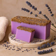 Two purple soap bars with a textured brown top on a beige surface with lavender flowers.