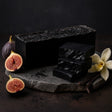 Black soap bars on a stone surface with figs and a flower on a dark background