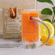 Orange soap bar with a rope, next to its packaging and citrus fruits on a neutral background