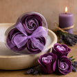 Purple soap flowers in a clear container with a purple candle and lavender on a wooden surface.