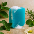 Blue bar of soap with foam surrounded by green leaves and a white flower on a wooden surface.
