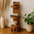 Wooden tiered shelf with books, a candle, and a bowl, accompanied by a vase with dried plants and a potted plant on a wooden floor.