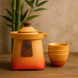 Orange ceramic teapot with wooden lid and matching bowls on a wooden surface with a bamboo plant in the background.